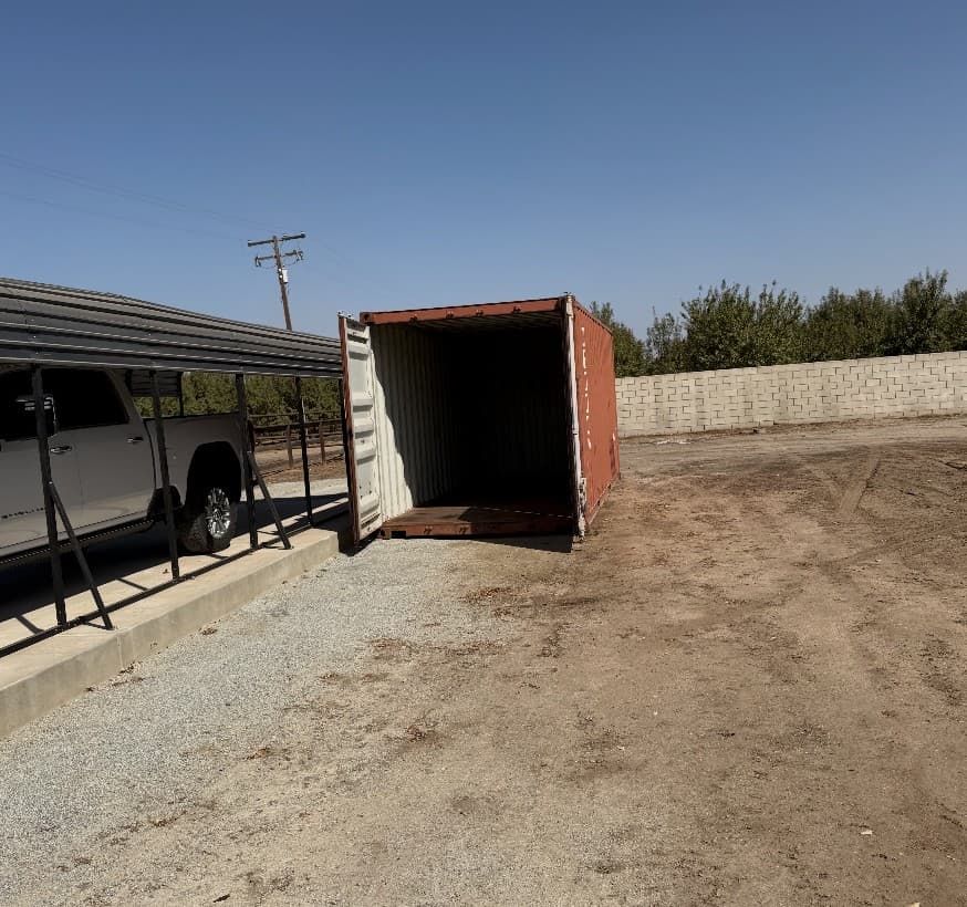 Used oxide-red shipping container with cargo doors open on a gravel pad next to a carport