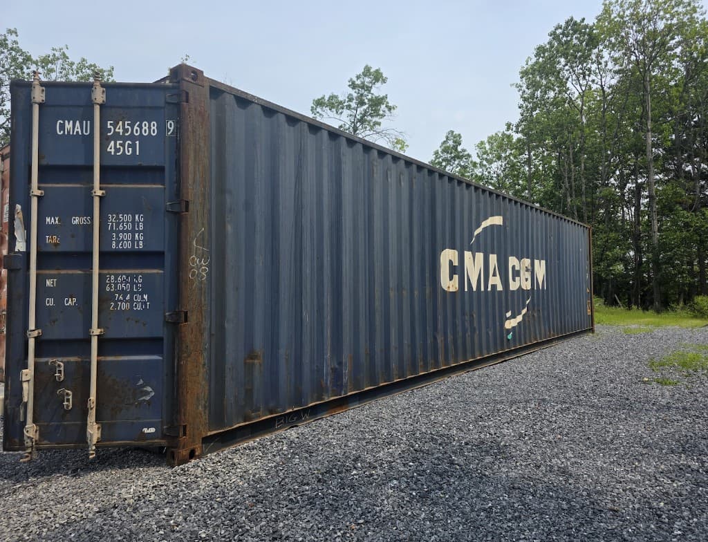 Side of a used blue CMA CGM 40ft shipping container on gravel, faded logo and rust along the bottom rail